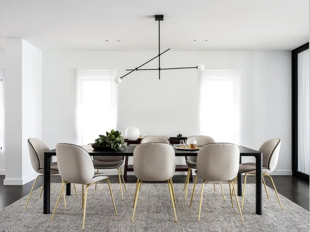Bright contemporary dining room with a long black rectangular dining table surrounded by upholstered curved-back chairs with slender gold-toned legs. White walls and sheer window coverings create an airy backdrop, while a sculptural black linear pendant light hangs above the table. Dark timber flooring, a large grey area rug, potted greenery, and minimal table styling complete the refined interior.