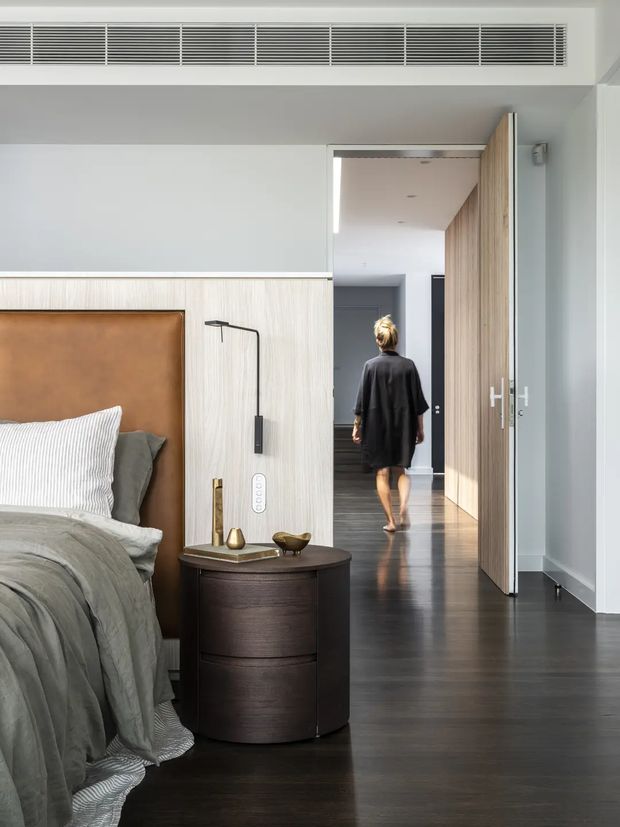 A refined contemporary bedroom with a partial view toward a hallway. The room features a large upholstered tan leather headboard set against pale vertical timber wall panelling, dark timber flooring, grey layered bedding, a rounded dark timber bedside table, and a slim black wall-mounted reading light. A full-height timber door opens to a bright corridor with matching timber wall detailing. The restrained palette and clean built-in detailing create a calm, sophisticated interior.