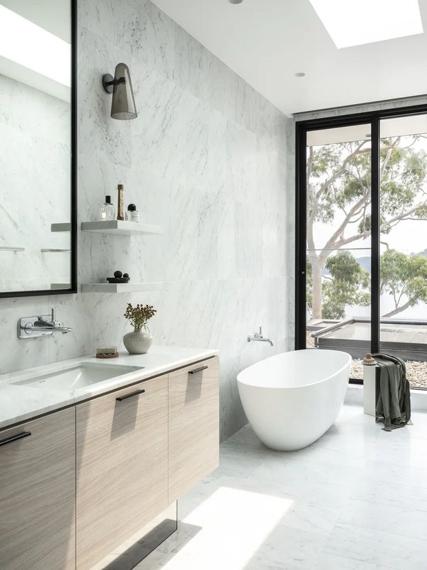Bright contemporary bathroom featuring full-height white marble-look wall and floor surfaces, a floating light-wood vanity with integrated white basin, wall-mounted dark tapware, open display shelves, a freestanding white oval bathtub, black-framed full-height glazing, and a skylight. The room overlooks trees and appears airy and refined.