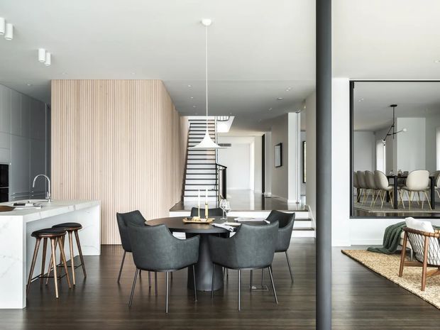 Contemporary open-plan interior combining a kitchen, dining area and living zone. The room features dark timber flooring, a white island with integrated sink, pale vertical timber slat screening, a round dark dining table with upholstered chairs, a sculptural pendant light, and a large interior window reflecting the dining space. A staircase and hallway extend toward the rear, creating a spacious, polished layout.