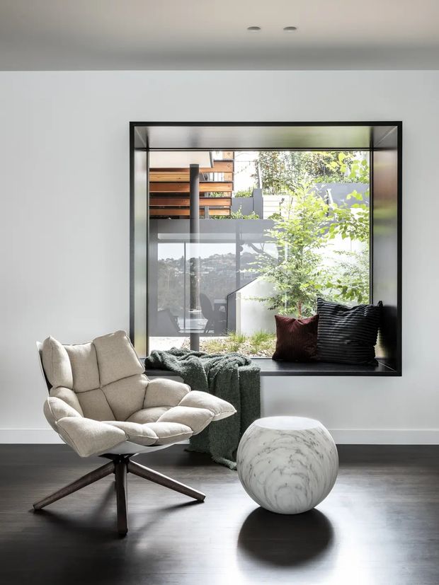 A minimalist sitting nook with a sculptural cream lounge chair on a dark timber swivel base, a round white marble-like side table, and a deep black-framed window seat. The window overlooks a contemporary multi-level exterior, while a textured green throw and dark cushions add softness and contrast. The room has smooth white walls, recessed ceiling lights, and dark polished timber flooring.