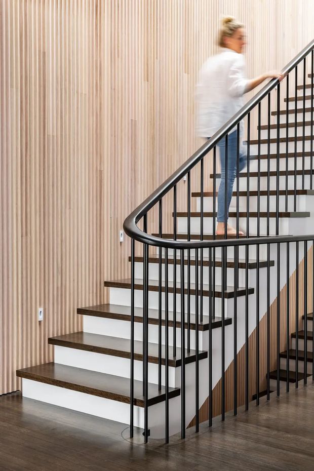 Interior view of a modern stairway with a sculptural curved black metal balustrade, dark timber stair treads, white stair risers, and a vertically slatted light timber feature wall. The staircase turns around a central void, with polished dark flooring and a blurred person ascending.