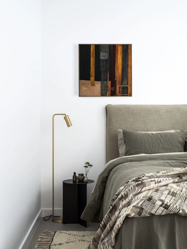 A softly styled bedroom corner with a large upholstered grey bed, layered grey bedding, a patterned throw, and a small black bedside table. A slim brass floor lamp stands beside the bed, while an abstract mixed-media artwork hangs on the white wall above. A neutral textured rug adds warmth to the otherwise restrained palette.