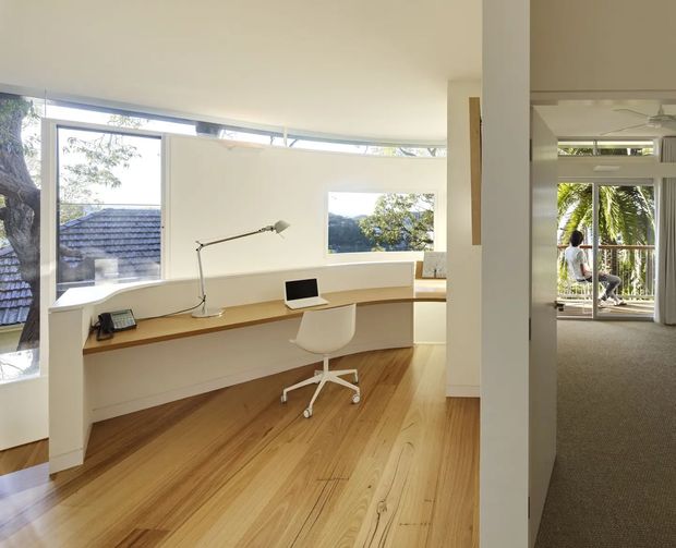 Bright open-plan home office with a curved built-in white desk and timber work surface positioned in front of expansive corner glazing. The room features warm timber flooring, white walls, a minimalist task lamp, computer, and ergonomic white chair. An adjoining carpeted living area is visible through the opening, with additional glazing and tropical greenery beyond.