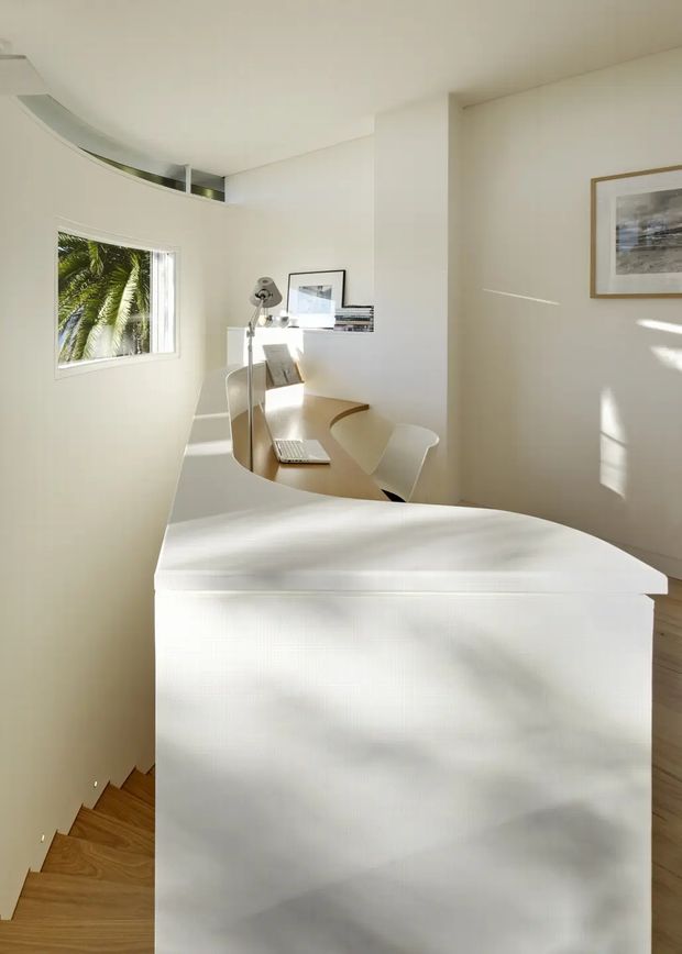 A bright, minimalist home office nook viewed from behind a curved white partition or built-in enclosure. The compact workspace includes a pale curved desk, white chair, laptop, desk lamp, framed artwork, and built-in shelving. White walls and joinery, warm timber flooring, abundant natural light, and a curved internal opening create a sculptural contemporary interior.