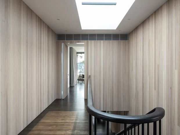 A narrow, refined hallway or stair landing lined with vertical light timber battens. A dark timber floor leads toward an open doorway, while a curved black handrail frames the stair opening in the foreground. A large rectangular skylight and linear ceiling ventilation grille provide overhead detail and natural light.
