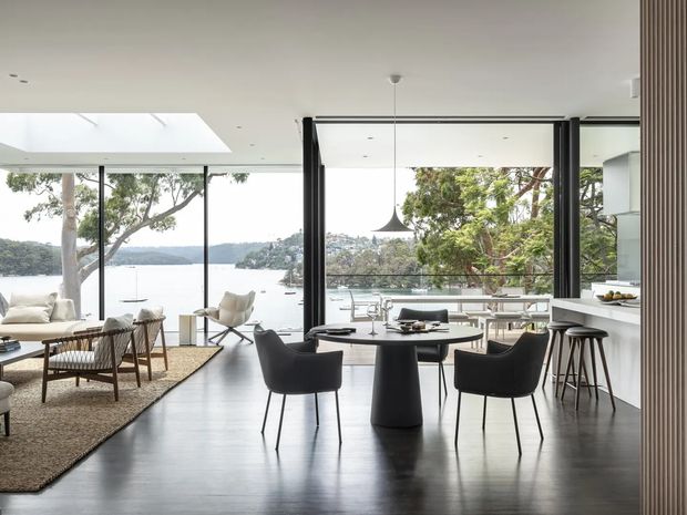 Open-plan contemporary interior combining a dining area, living area and kitchen, with floor-to-ceiling black-framed glazing overlooking a body of water. The space has dark timber flooring, a sculptural round dining table with upholstered chairs, a neutral lounge setting, minimalist white cabinetry, a pendant light and a large skylight.