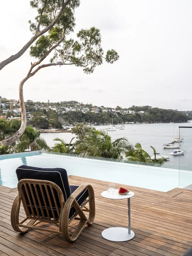A bright waterfront outdoor lounge overlooking a calm harbour, with an infinity-edge pool, timber decking, tropical planting, a woven timber lounge chair, and a small round side table. Boats and a leafy residential shoreline are visible beyond the frameless glass pool barrier. The scene has a relaxed, resort-like contemporary feel.