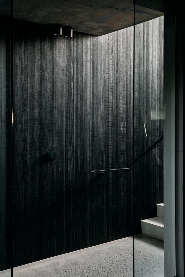 A moody, minimalist interior stairwell viewed through a frameless glass balustrade. The space features dramatic dark vertically textured wall cladding, a raw concrete ceiling, polished grey concrete flooring, exposed concrete stair treads, and a slim black metal handrail. Sparse recessed or pendant-style lighting emphasizes the industrial, sculptural character.