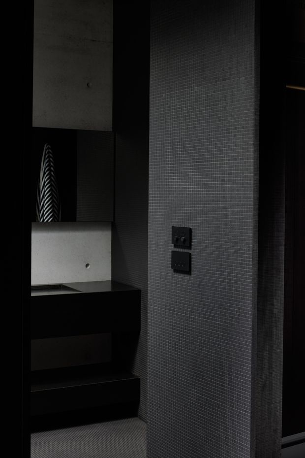 A dark, highly minimalist interior vignette featuring a black built-in console with drawers, a pale concrete-like rectangular wall feature, and a small sculptural object. The central wall is clad in tiny dark grey grid-pattern tiles, with black electrical outlets and deep black surrounding surfaces. The composition is moody, refined, and strongly monochromatic, with crisp geometric lines and subtle texture.