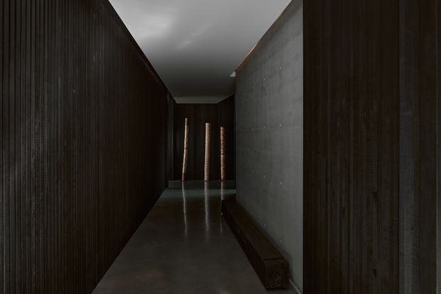 A dark, narrow architectural corridor with heavily ribbed dark timber wall cladding on both sides, a raw grey concrete wall, and a highly polished dark floor reflecting the walls and slatted openings at the far end. The space has a stark, minimalist and dramatic character with subdued natural light.