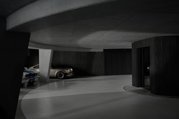 A dark, sculptural covered vehicle forecourt or garage with a broad curved concrete ceiling, smooth concrete floor, black ribbed wall cladding, and angular structural columns. Two cars are parked in recessed bays, while a dark entry door is set into the ribbed enclosure on the right. The architecture has a dramatic, minimalist monolithic quality.