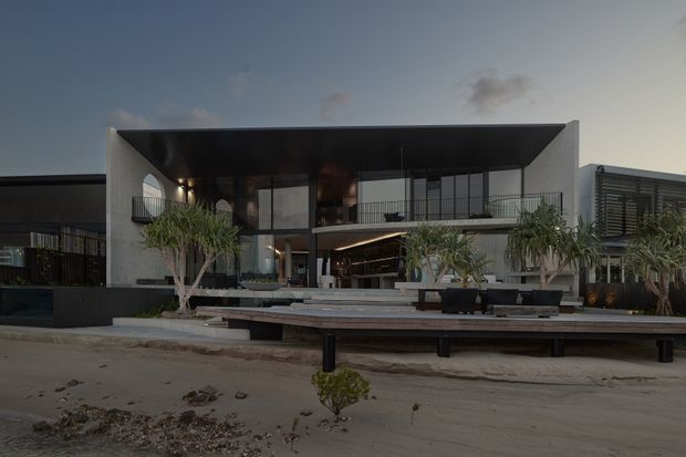 Contemporary multi-level beachfront or waterfront residence viewed from the exterior at dusk. The house features a strong flat-roofed geometric form, deep projecting roof canopy, extensive floor-to-ceiling glazing, dark metal framing, pale rendered walls, upper-level terraces with metal balustrades, and landscaped planters. A broad timber deck or boardwalk and sandy foreground reinforce the coastal setting.