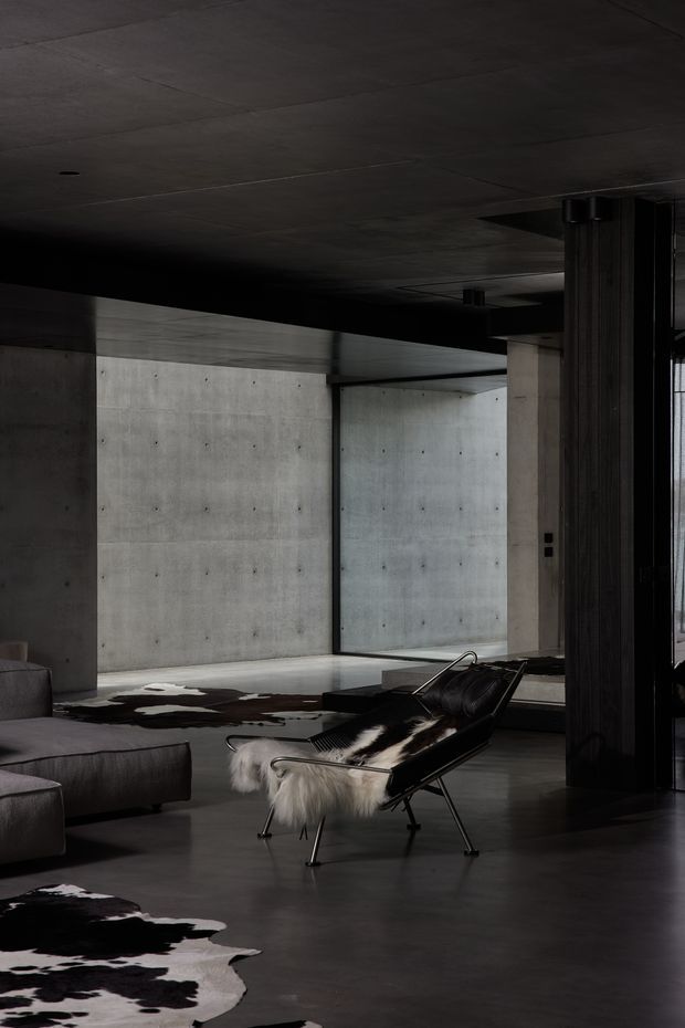 A dramatic, moody modern living room with exposed concrete walls and ceiling, dark polished flooring, broad open circulation, and full-height glazing or openings. A large grey sectional sofa and sculptural black lounge chair with a white fur throw sit over black-and-white cowhide rugs. Dark timber or black architectural columns and beams create a strong structural frame.