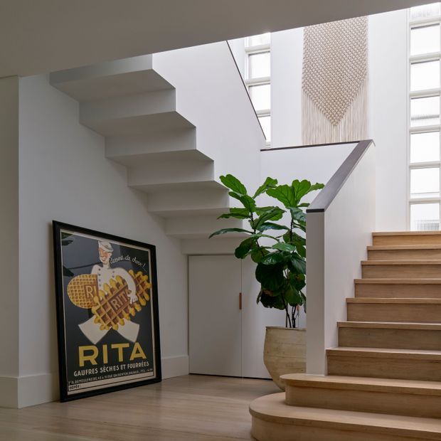 Bright contemporary stair hall with white walls, a sculptural open-riser staircase, warm timber treads and flooring, tall glazed openings, a large potted plant, and framed artwork. A second timber staircase rises along the right side, creating a layered vertical circulation space with abundant natural light.