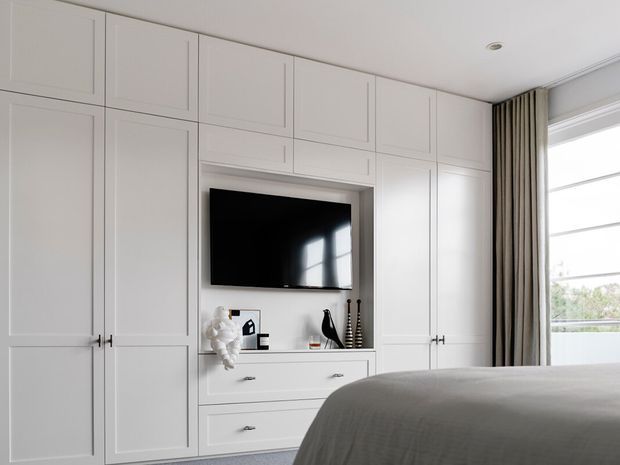 A bright, refined bedroom with full-height white built-in cabinetry spanning the wall, including paneled wardrobe doors, drawers, open display shelving, and a wall-mounted television. A large bed with grey bedding occupies the foreground, while floor-to-ceiling curtains and a large window provide natural light.