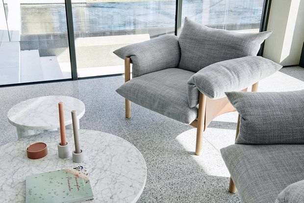 Bright contemporary living room with two sculptural grey upholstered lounge chairs, pale timber legs, round white marble coffee tables, and a speckled grey carpet. Full-height black-framed glazing opens the room visually to an exterior terrace or balcony, with strong natural daylight and a restrained neutral palette.