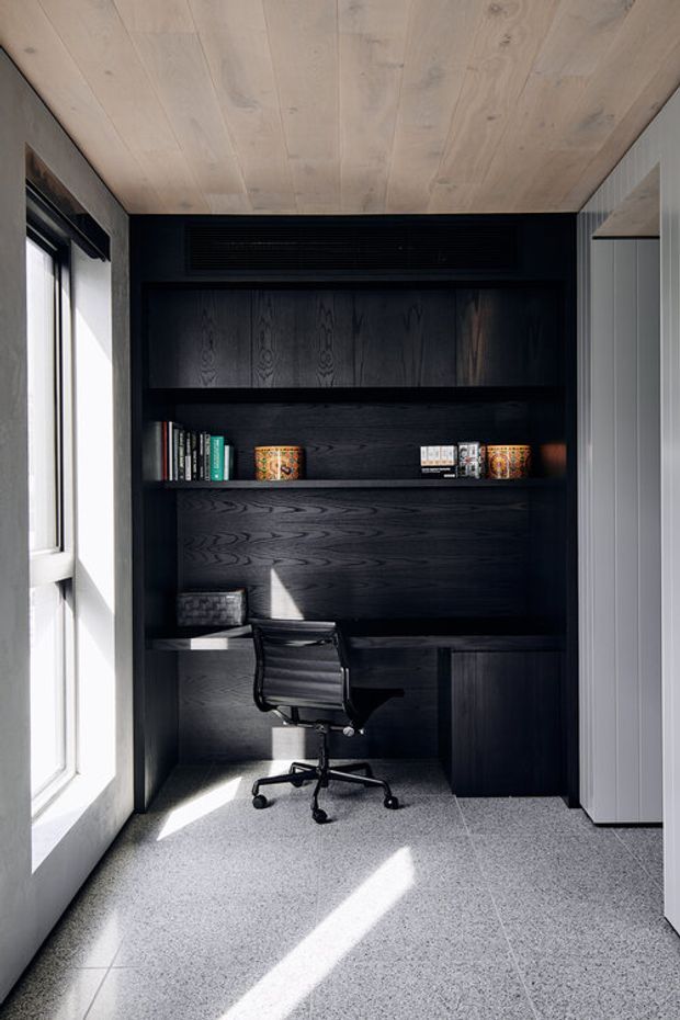 Compact, contemporary home office nook with a built-in black-stained timber desk, shelving, cabinetry and integrated ventilation. A black ergonomic office chair sits on a light grey carpet, with tall glazing on the left bringing in strong daylight. The ceiling is clad in pale timber boards, while the adjacent wall has light vertical cabinetry or panelling.