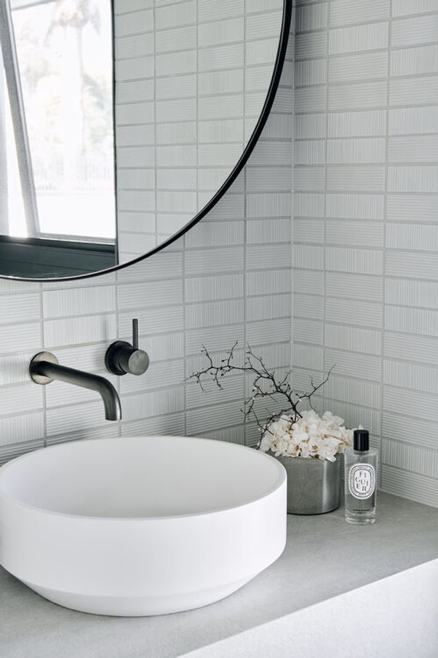 Close-up of a refined contemporary bathroom vanity with a white vessel basin on a light grey benchtop, brushed dark metal wall-mounted tapware, a large round mirror, and pale linear textured wall tiles. Minimal styling includes a small vase of branches and a fragrance bottle.