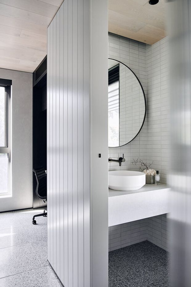 A contemporary bathroom viewed through a partially open doorway, featuring a floating pale vanity with a white vessel basin, wall-mounted black tapware, a large round black-framed mirror, and white horizontally stacked wall tiles. The space has a light timber-lined ceiling, vertically grooved white wall panelling at the doorway, and dark terrazzo-style flooring. An adjacent compact office or workspace is visible to the left.