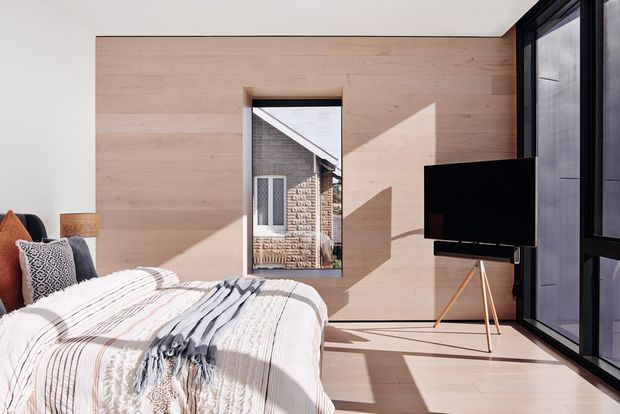 Bright contemporary bedroom with pale timber wall panelling and matching timber flooring. A bed with layered white and grey striped bedding occupies the foreground, while a large black television on a slim tripod stand sits beside full-height dark-framed glazing. A narrow window reflects or reveals a textured masonry exterior, and strong sunlight creates geometric shadows across the room.
