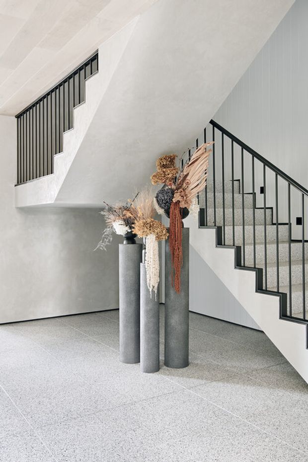 A minimalist interior stair hall with two intersecting flights of stairs, black metal balustrades, pale textured walls, and a light terrazzo floor. Three tall cylindrical grey concrete or stone plinths display dried botanical arrangements beneath the stairs.