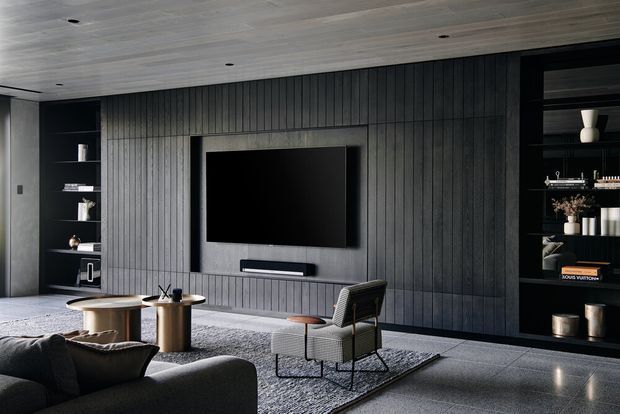 A sophisticated contemporary living room with a large wall-mounted television set into a dark vertically panelled feature wall. Built-in black shelving displays books and decorative objects on either side. The room has a pale timber-clad ceiling, polished grey floor, textured grey rug, low coffee tables, a lounge chair, and part of a dark sofa in the foreground.