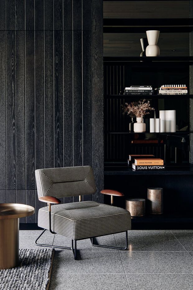 A moody, refined interior vignette featuring a black timber-paneled wall, built-in black shelving with curated books and ceramic vessels, and a mid-century-inspired upholstered lounge chair with wooden armrests. A small round side table, textured rug, dark wood table, and grey tiled floor complete the sophisticated setting.