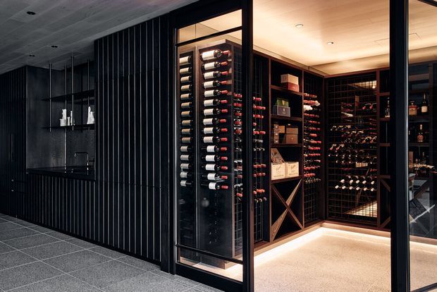 A sophisticated walk-in wine room viewed from an adjacent dark-toned interior. Floor-to-ceiling black-framed glass doors reveal extensive wine bottle racking, timber-toned cabinetry, display shelving, and a softly illuminated pale carpeted floor. The surrounding space features charcoal vertical wall panelling, dark cabinetry, and grey tiled flooring, creating a moody, refined atmosphere.