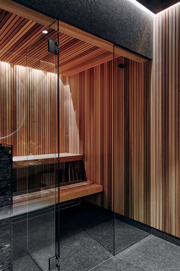 A refined compact sauna or steam-room interior enclosed by clear glass, with richly stained vertical timber slat walls and matching slatted ceiling. Built-in timber benches line the walls, while dark charcoal stone-look floor tiles, black trim, and discreet integrated lighting create a warm, moody, contemporary atmosphere.