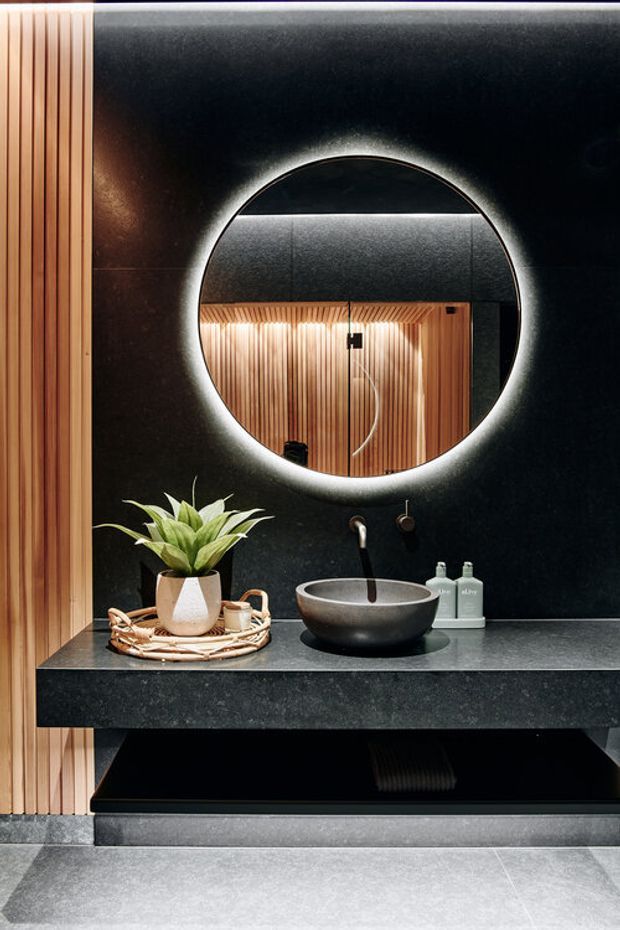 A sophisticated contemporary bathroom vanity featuring a round backlit mirror, matte black wall finish, dark stone benchtop, vessel basin with wall-mounted tapware, and warm vertical timber battens. A floating black vanity provides open storage below, while a small plant and toiletries add subtle styling.
