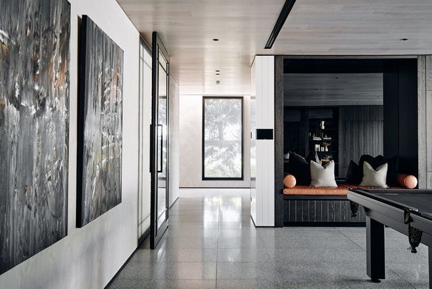 A refined contemporary interior hallway opening into a dark built-in lounge or media nook. Large abstract artwork lines the pale wall, while full-height glazing and a central window bring in natural light. The space features a polished grey tiled floor, light timber ceiling, dark timber wall panelling, integrated bench seating with cushions, and a partially visible pool table in the foreground.