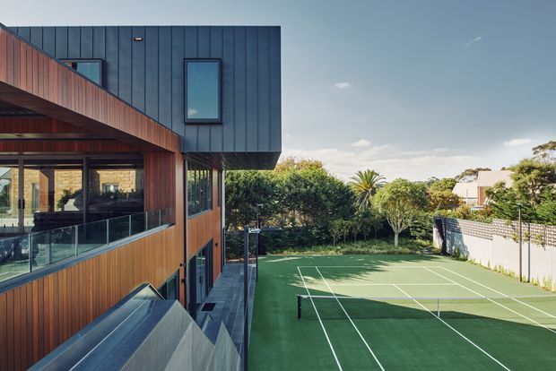 Exterior view of a contemporary multi-level residence with dark vertical metal cladding, warm timber wall cladding, extensive glazing and glass balustrades. A full-size green tennis court occupies the foreground beside landscaped gardens and neighboring homes.