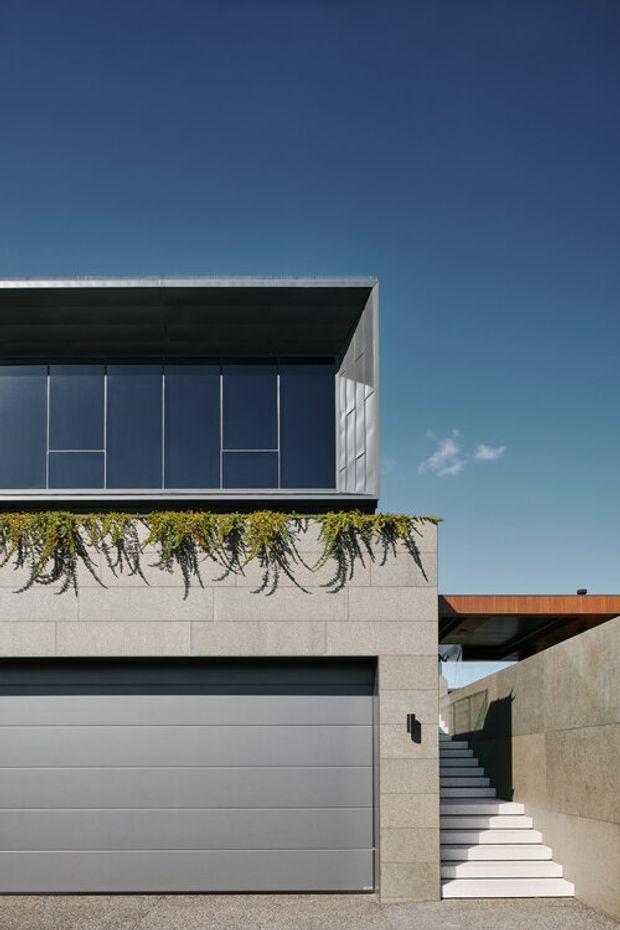 Exterior view of a contemporary multi-level residence with a large grey sectional garage door, pale stone or concrete-clad walls, a broad upper-level window, rooftop planting spilling over a parapet, and an external stair leading beneath a timber-toned flat-roof canopy. The composition is sharply geometric with strong horizontal and vertical lines against a clear blue sky.