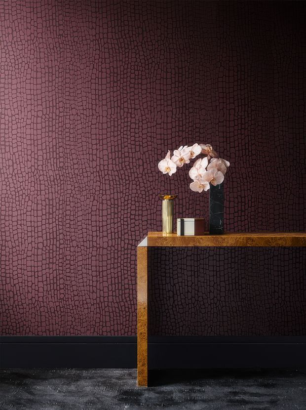 A styled interior vignette featuring a slim console table with a warm metallic gold finish against a dramatic dark burgundy textured wall. The table holds a tall gold vessel, small white container, and a black marble vase with pale orchid stems. A black skirting board and dark textured floor reinforce the moody, luxurious palette.