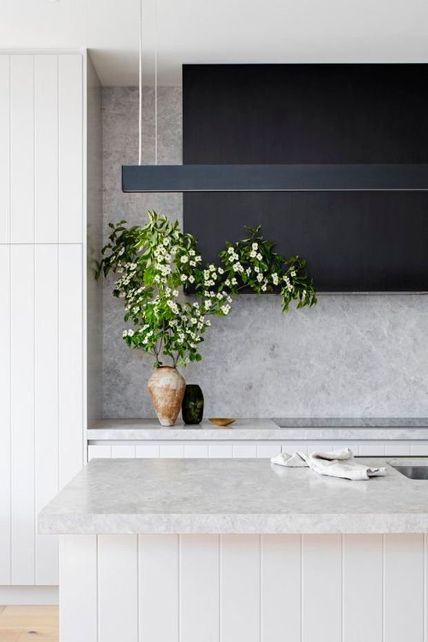 Bright contemporary kitchen featuring white vertical-panel cabinetry, a pale stone island and benchtop, a textured grey stone splashback, and a large dark feature panel behind a suspended linear pendant light. A floral arrangement and simple decorative vessels add a natural accent.