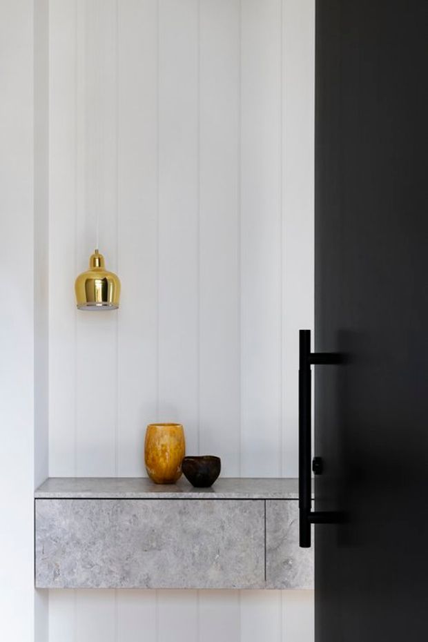 A refined minimalist interior vignette featuring a wall-mounted grey stone console with matching stone benchtop, set against white vertical wall panelling. A black pivot door with a bold black handle occupies the right side, while a small brass pendant light and simple ceramic vessels provide restrained decorative accents.