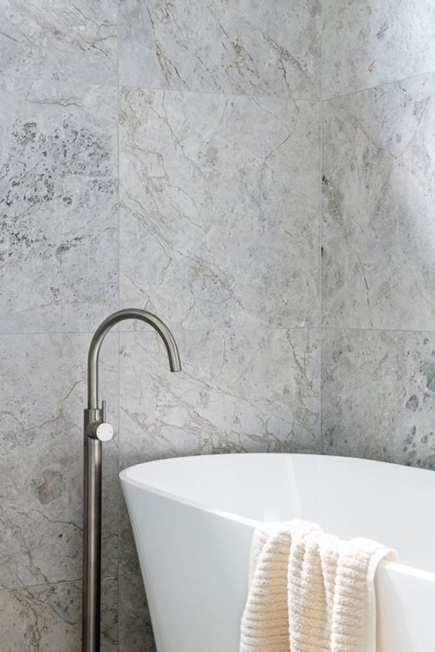 Close-up of a contemporary bathroom featuring a freestanding white bathtub, a tall curved brushed metal bath tap, and large-format pale grey natural stone-look wall tiles with veined detailing. A textured cream towel hangs over the tub edge.