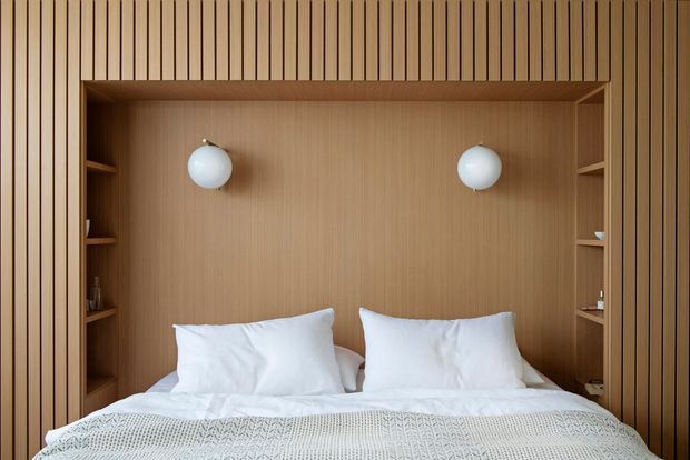 A warm, compact bedroom with a built-in timber wall alcove framing a double bed. Vertical timber battens continue across the wall and integrated side shelving, while two spherical wall lights flank the bed. The bed is dressed in white linens with a textured light throw, creating a calm, refined minimalist interior.
