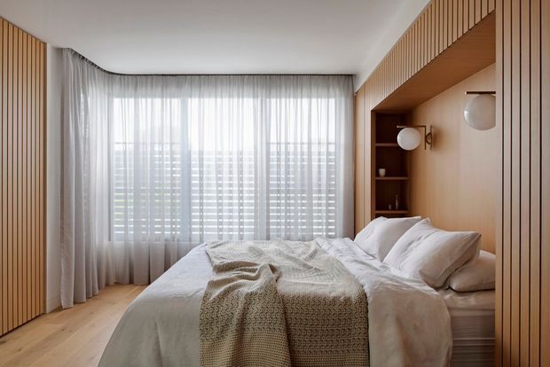 A warm, minimalist bedroom with a low-profile bed dressed in white linens and textured beige throws. Full-height sheer curtains cover a broad window, while light timber wall panelling, integrated shelving, overhead cabinetry, and spherical wall lights create a cohesive built-in joinery treatment.