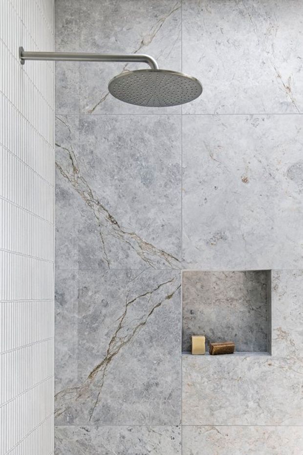 Close-up view of a contemporary walk-in shower featuring large grey marble-look wall tiles with prominent veining, a recessed tiled niche, a round brushed metal rain showerhead, and a narrow vertically ribbed tile accent wall.