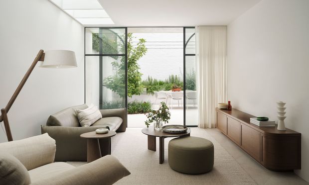 A bright, refined living room with a neutral palette, soft upholstered seating, a round coffee table and ottoman, and a low timber media console. Full-height black-framed glazing opens to a planted outdoor courtyard, while a roof skylight brings in additional natural light. The room has a calm, contemporary minimalist character with warm wood accents.