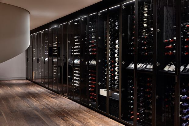 A dark, sophisticated wine storage room lined with tall black-framed glass-fronted wine racks and cabinets filled with bottles. The space has warm timber flooring, subtle integrated lighting, and a curved light-coloured wall at the end, creating a refined cellar-like atmosphere.