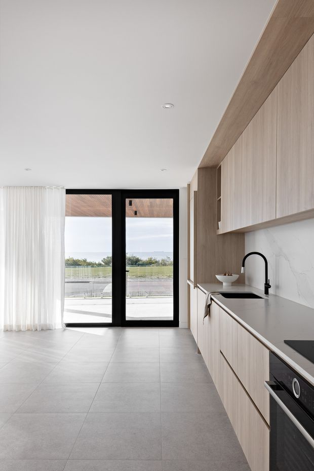 A bright, contemporary kitchen with a long run of light timber cabinetry, a dark grey benchtop, white marble-look splashback, black tapware and integrated appliances. Large black-framed glazed doors open to an outdoor terrace, while sheer white curtains and large-format grey floor tiles create a clean, refined atmosphere.