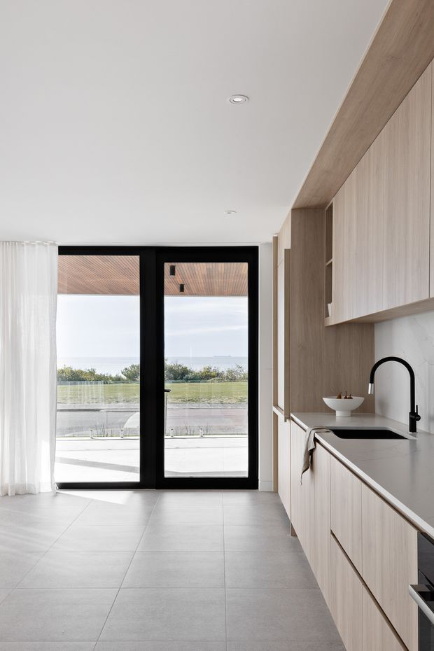 Bright contemporary kitchen with pale timber built-in cabinetry, a white stone benchtop and splashback, black-framed glazed doors, and large-format light grey floor tiles. The kitchen opens toward an outdoor terrace and landscaped outlook, with sheer curtains and abundant natural light.