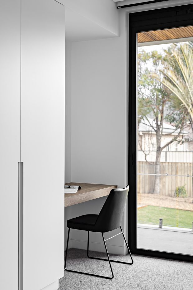 A minimalist home office nook beside a large black-framed glazed door or window. A slim timber desk is built into the corner, paired with a black upholstered chair, while white walls and cabinetry and a light grey carpet create a restrained monochromatic interior. The glazing looks onto a small garden and covered outdoor area.