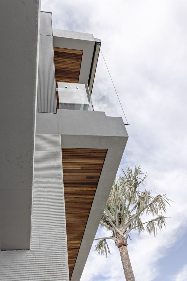 Upward-looking exterior view of a contemporary multi-level building with projecting concrete balconies or overhangs, warm timber soffit lining, corrugated light-grey cladding, and a clear glass balustrade. A palm tree is visible beside the structure against an overcast sky.
