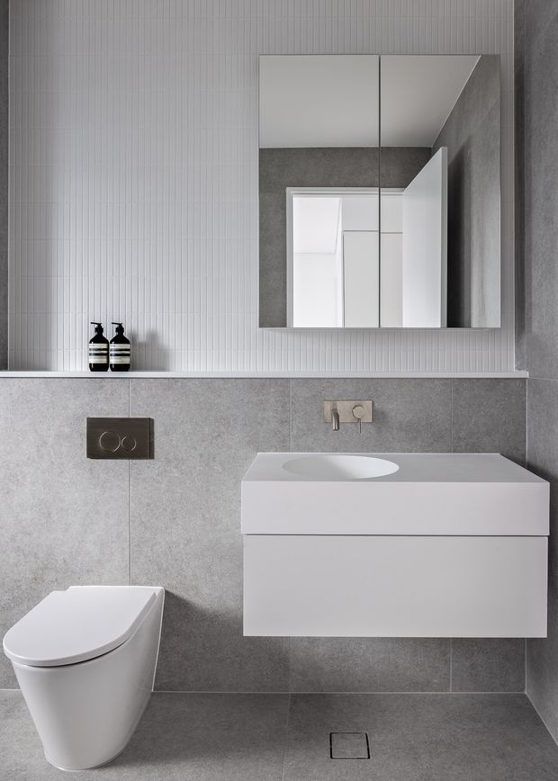 Contemporary compact bathroom in a monochromatic grey and white palette. The room features large-format grey wall and floor tiles, a white floating vanity with integrated basin, wall-mounted tapware, a wall-hung toilet, a dark flush plate, and a mirrored shaving cabinet. A vertically ribbed white wall surface adds subtle texture above the tiled dado.