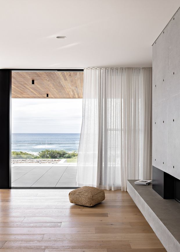 A minimalist contemporary living room opening onto a coastal terrace, with expansive black-framed glazing, sheer white curtains, warm timber flooring, and a textured concrete feature wall incorporating a long built-in fireplace or hearth. The ocean and beach are visible beyond the terrace. A woven floor cushion and a small stack of books provide understated furnishings.