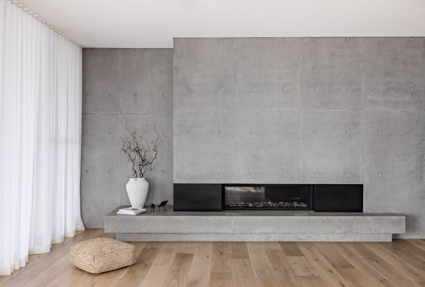 Minimalist living room with a full-height exposed concrete feature wall and a long built-in linear fireplace set into dark cabinetry. The room has light timber flooring, sheer white curtains, a simple concrete hearth, and sparse neutral decor including a woven floor cushion and ceramic vase.